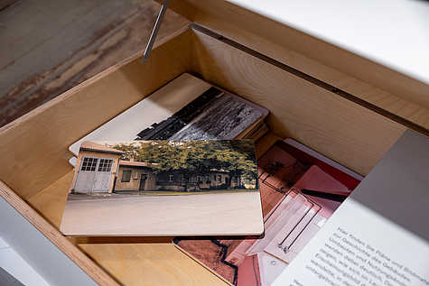 Innenansicht einer Ausstellung mit einer Holzkiste, die Postkarten und Bilder enthält. Eine Postkarte zeigt ein altes Gebäude vor Bäumen.